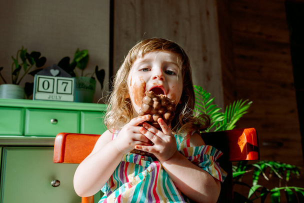 Little girl is dirty with chocolate. The girl smeared chocolate, dirty hands and face. Sweet fingers, lips and cheeks. World Chocolate Day July 7th. - Foto, imagen