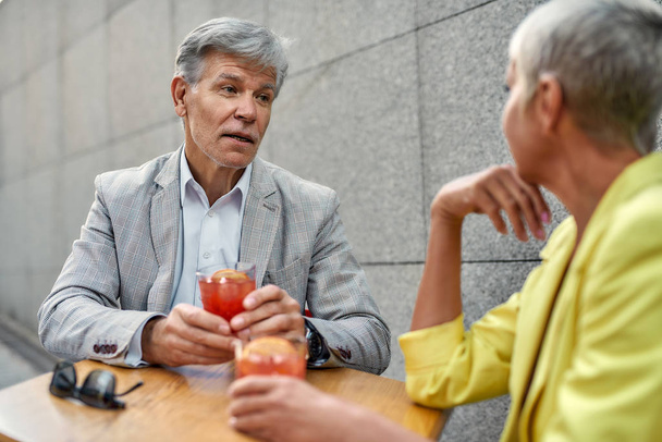 Schöner reifer Mann in formeller Kleidung, der Cocktail hält und etwas mit seiner Freundin diskutiert, während er zusammen im Café im Freien sitzt - Foto, Bild