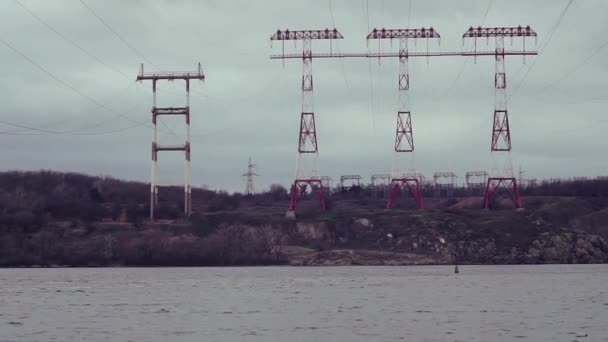 3 Power lines with crane-white foundations on the other side river, rocks. Spring, winter, autumn, cold - Video