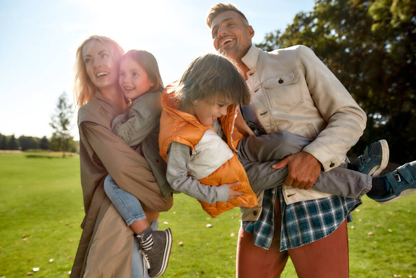 Гра на відкритому повітрі. Молоді батьки та їхні діти насолоджуються сонячним днем і розважаються разом, стоячи на зеленому лузі
 - Фото, зображення
