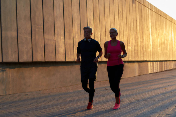 Νοιαζόμαστε για την υγεία μας. Ενεργό ζευγάρι ηλικιωμένων σε αθλητικά ρούχα τρέχει μαζί μέσα από το δρόμο της πόλης το πρωί - Φωτογραφία, εικόνα