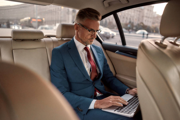 Effective business strategy. Confident mature businessman in full suit working on his laptop while sitting in the car - Fotoğraf, Görsel