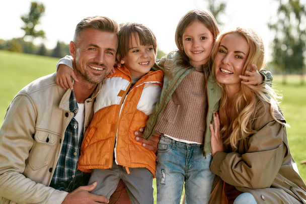 Relajándonos juntos. Retrato de feliz hermosa familia con los niños abrazando y mirando a la cámara con sonrisa mientras pasa tiempo al aire libre
 - Foto, imagen