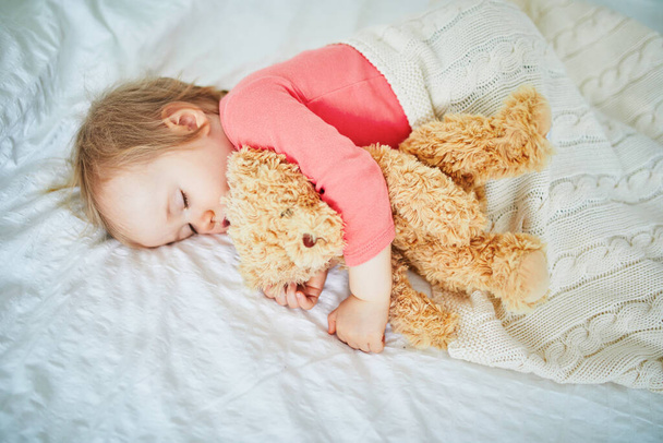 Adorable baby girl sleeping with her favorite toy. Little child having a day nap and hugging a teddy bear. Infant kid resting in nursery - Фото, изображение