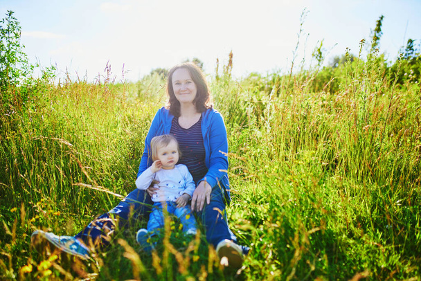 Чарівна дівчинка сидить на зеленому полі зі своєю матір'ю. Молода жінка і її дочка розважаються на відкритому повітрі. Малюк досліджує природу
 - Фото, зображення
