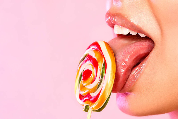 una joven hermosa lame una colorida y brillante piruleta fotografiada en primer plano con su lengua. sobre un fondo rosa aislado. con copyspace
 - Foto, imagen