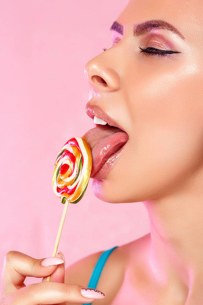 a young beautiful girl licks a colorful bright Lollipop photographed in close-up with her tongue. on a pink isolated background. with copyspace - Photo, image
