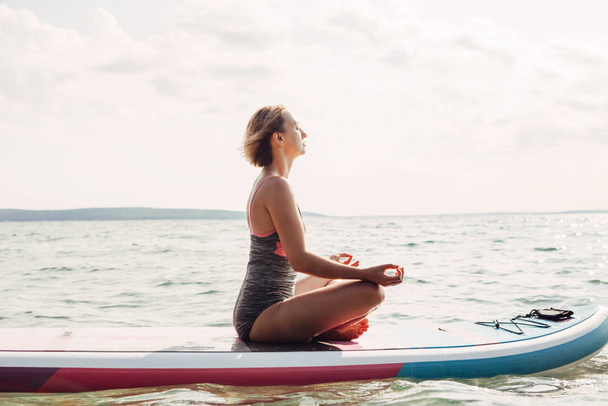 Jonge blanke vrouw die yoga beoefent op paddle sup surfplank bij zonsondergang. Vrouwelijke stretching doen workout op meer water. Moderne individuele hipster outdoor seizoensgebonden zomersport activiteit. - Foto, afbeelding