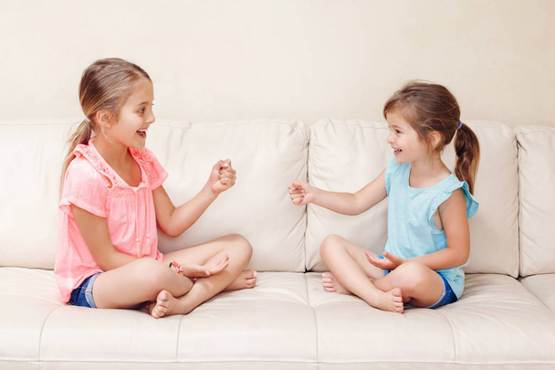 Kaksi tyttöä ystäviä pelaa kivi paperi sakset käsi peli. Valkoihoiset lapset istuvat sohvalla leikkimässä yhdessä. Mielenkiintoista viihdyttävää toimintaa lapsille. Aito vilpitön elämäntapa hetki.  - Valokuva, kuva