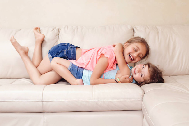 Two cute little Caucasian girls siblings playing at home. Adorable children kids lying hugging on couch together. Authentic candid lifestyle domestic life moment. Happy friends sisters relationship.  - Foto, Imagem