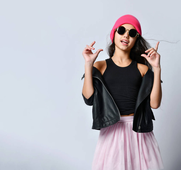Teenage female in black leather jacket pink hat and sunglasses. She is gesticulating while posing isolated on white. Close up - Fotoğraf, Görsel