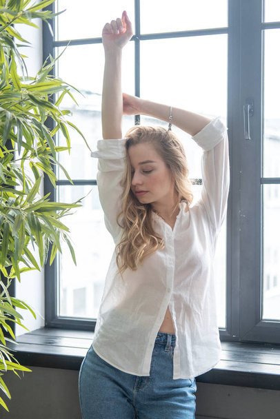 Young beutiful blond woman wearing white shirt and blue jeans standing next to window. Morning portrait of a girl with window and green plants on the background - Fotografie, Obrázek