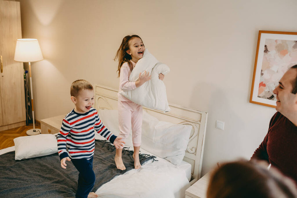 Linda menina caucasiana segurando um travesseiro e brincando com seu irmão mais novo, enquanto seus pais estão assistindo-los
 - Foto, Imagem