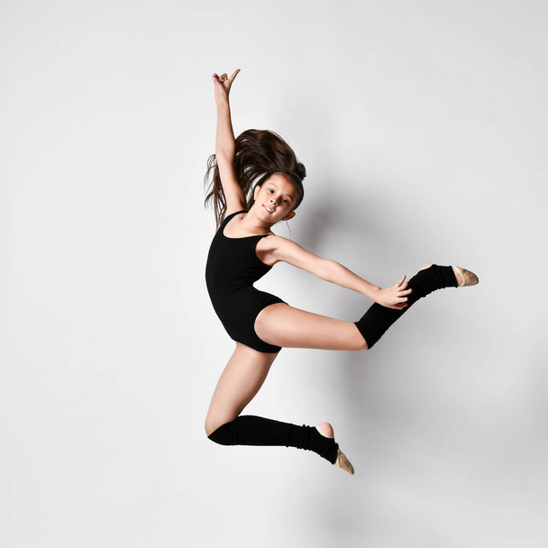 Teenage brunette girl gymnast with pigtail, in black leotard, knee socks and ballet shoes, performing exercises, posing isolated on white. Close-up. - Foto, imagen
