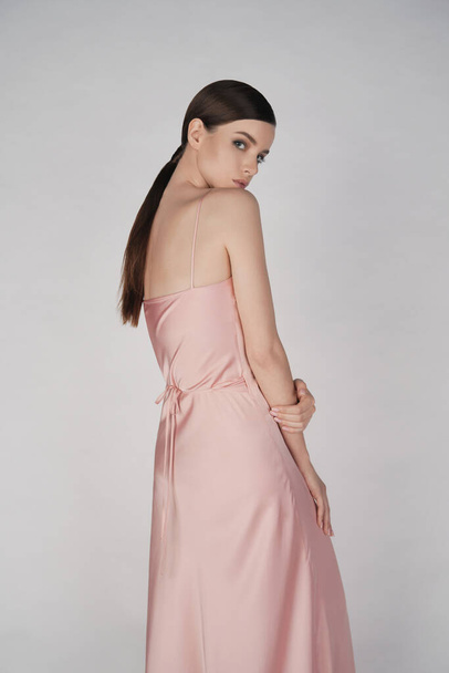 Woman in elegant fashionable dress. Beautiful model pose in studio in pink evening clothes. Classic silk dress. Sexy lady stay on white background. Perfect young woman with classic hairstyle and makeup.  - Foto, Imagen