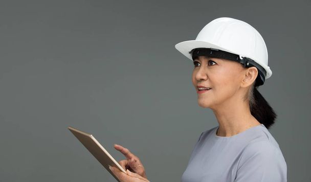 Beautiful Asian Smart Architect Engineer 50s 60s years old woman in white hard hat helmet hold tablet iot, portrait studio lighting gray background copy space, concept aging society woman can do - Zdjęcie, obraz