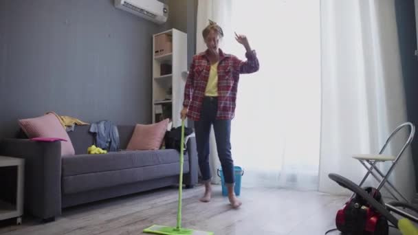 Happy young woman cute housewife is washing floor with mop and dancing at home - Filmmaterial, Video