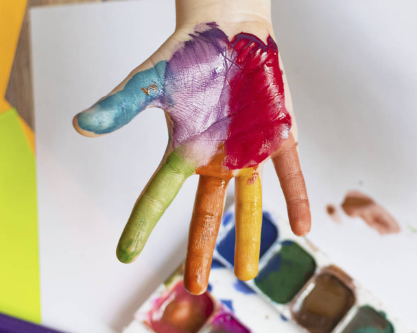The child paints. Palms and hands of a child in multi-colored watercolor. Watercolor, gouache, paint brushes, colored and wax crayons, stickers. Set for drawing, creativity and hobbies - Photo, Image