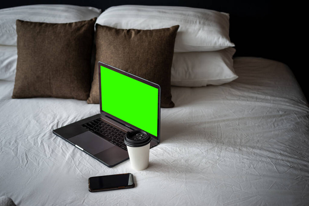 Coffee Cup To Go, Laptop and smartphone on bed. Working from home concept. Green template on screen. - Photo, Image