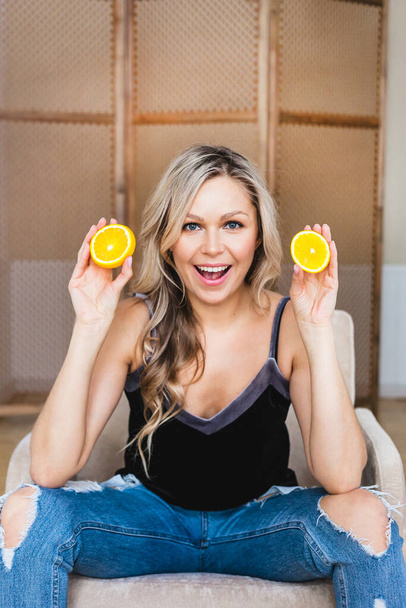  girl oEuropean appearance with blond hair stands near the mirror, girl's reflection mirror, holding cut orange, fruit, covering  eyes orange, vitamins, blouse, body care, makeup, morning treatments - Photo, Image