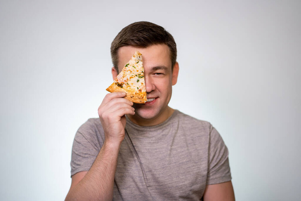 Man holding piece of pizza in front of their face. On a white background in a gray t-shirt. Quick, harmful, Italian food. - Foto, immagini