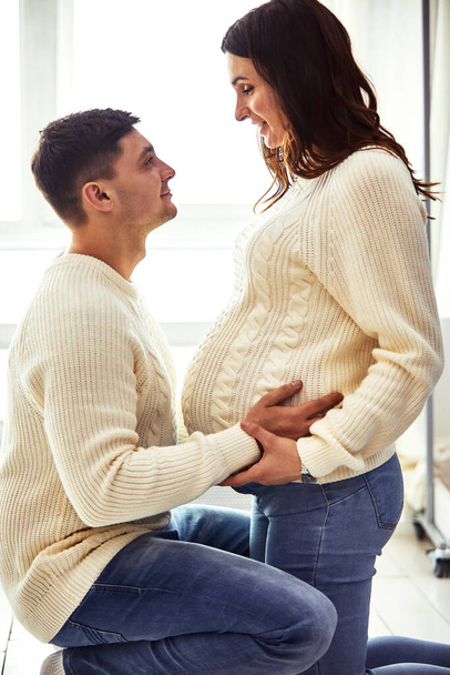Close up portrait of expecting teenage couple laughing happily, embracing baby in belly together. Couple dressed in blue and white colors. - Foto, Imagen