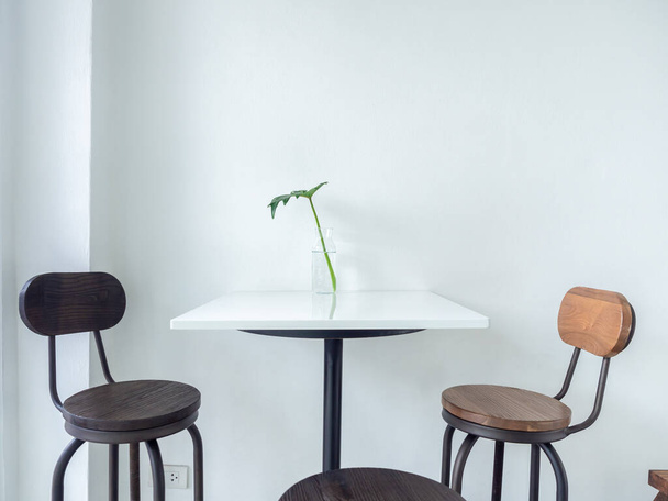Witte cafe decoratie minimale stijl. Glazen fles met tropisch palmblad op witte tafel en houten kruk bar op witte muur achtergrond met ruimte. - Foto, afbeelding