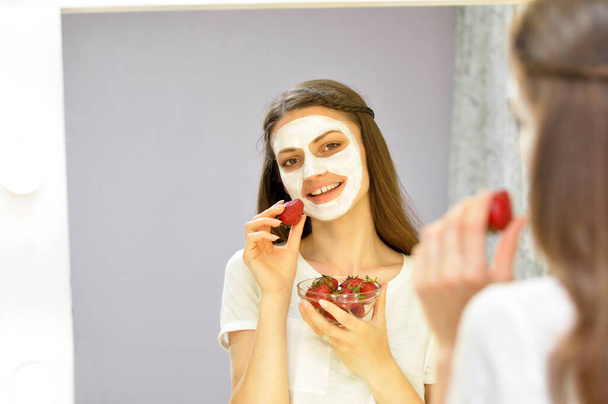 Μια νεαρή όμορφη γυναίκα με μάσκα κοσμετολογίας στο πρόσωπό της τρώει φράουλες και κοιτά στον καθρέφτη. - Φωτογραφία, εικόνα