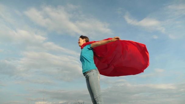 niño juega en abrigo rojo, sueño de convertirse en un héroe Superwoman. hermosa chica superhéroe de pie en el campo en una capa roja, capa revoloteando en el viento. En cámara lenta. chica sueña con convertirse en un superhéroe
. - Foto, Imagen