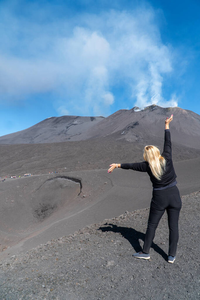 Sarı saçlı genç bir kadın Etna 'nın büyük volkanik kraterine karşı duruyor, kollarını kaldırıyor ve dumanlı volkana ve mavi gökyüzüne bakıyor. Arkadan turist fotoğrafı. İtalya, Catania (dizi)) - Fotoğraf, Görsel