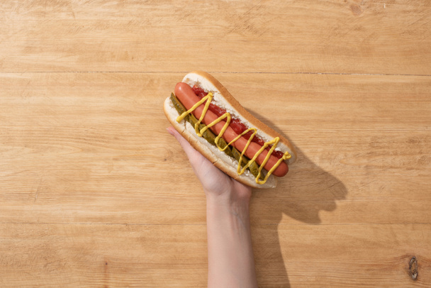 cropped view of woman holding delicious hot dog with pickles, mustard and ketchup on wooden table - Fotó, kép