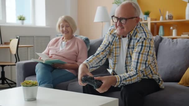 Slow motion of cheerful old man playing video game while woman reading book at home - Video