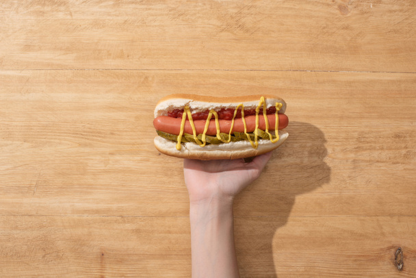 partial view of woman holding delicious hot dog with pickles, mustard and ketchup on wooden table - Photo, Image