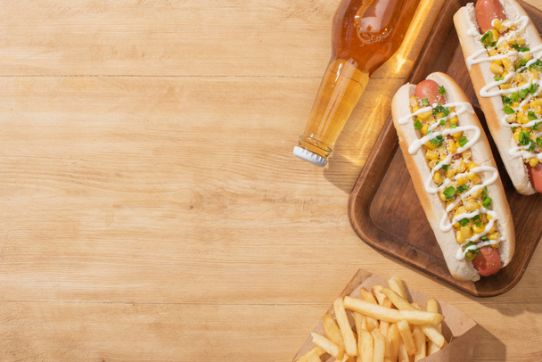 top view of delicious hot dogs with corn, green onion and mayonnaise near french fries and beer on wooden table - Photo, Image