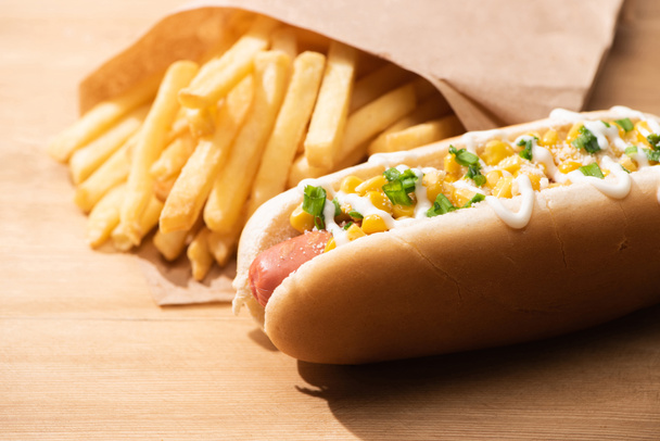 selective focus of delicious hot dog with corn, green onion and mayonnaise near french fries on wooden table - Zdjęcie, obraz