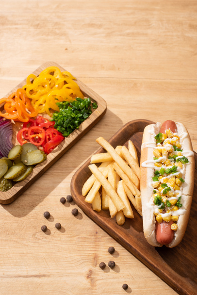 delicious hot dog near board with sliced vegetable and french fries on wooden table - Valokuva, kuva