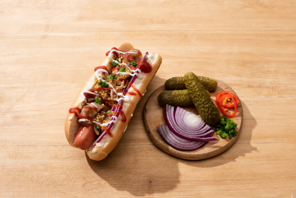 delicious hot dog with red onion, bacon and Dijon mustard near board with vegetables on wooden table - Photo, Image