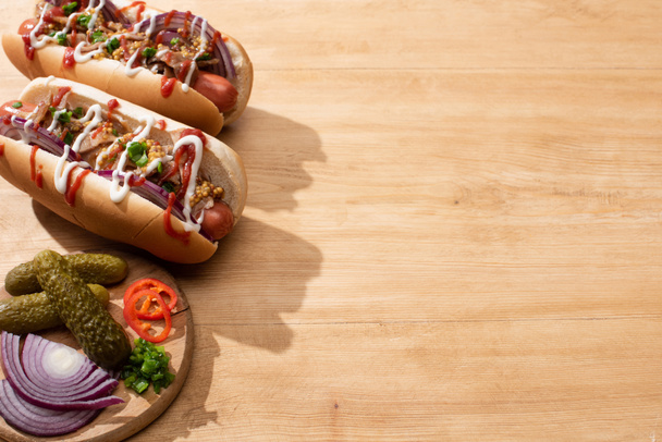 delicious hot dogs with red onion, bacon and Dijon mustard near board with vegetables on wooden table - Photo, Image