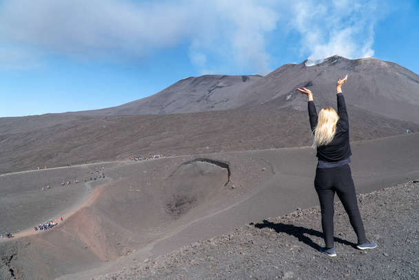 Eine junge Frau mit blonden Haaren steht vor dem großen Vulkankrater des Ätna, streckt die Arme in die Höhe und starrt auf den rauchigen Vulkan und den blauen Himmel. Touristenfoto von hinten. Sizilien, Italien, Catania (Serie)) - Foto, Bild