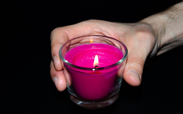 Яскраво-рожева свічка в скляній банці на чорному тлі. Свічка горить. Збережено. Рука підпалює свічку сірниками. Викладіть свічку пальцями, руками
. - Фото, зображення