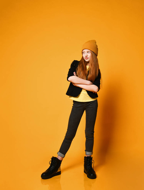 Cool red-haired girl teenager in hat black jacket tight jeans and boots is standing with her arms crossed over chest - 写真・画像