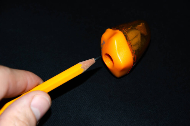 Рука тримає олівець і заточується в точилці. Олівець для олівця на чорному тлі. Помаранчевий олівець, помаранчева зміна, стружка
 - Фото, зображення