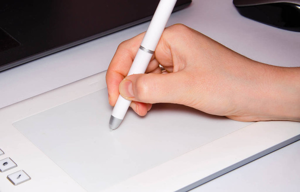 Vrouwelijke handen werken op een grafische tablet. Hand houdt stylus pen vast en tekent. Witte grafische tablet. Het werk van een grafisch ontwerper. Meisje werkt op een tablet aangesloten op een laptop. Achteraanzicht van achteren - Foto, afbeelding