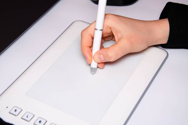 Vrouwelijke handen werken op een grafische tablet. Hand houdt stylus pen vast en tekent. Witte grafische tablet. Het werk van een grafisch ontwerper. Meisje werkt op een tablet aangesloten op een laptop. Achteraanzicht van achteren - Foto, afbeelding