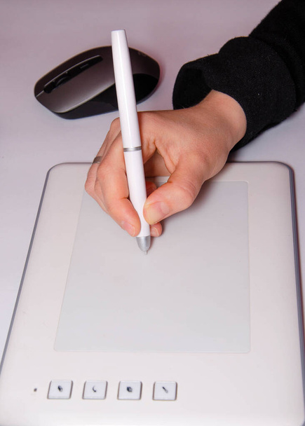 A női kezek grafikus táblán dolgoznak. A kéz fogja a tollat és rajzol. Fehér grafikus tabletta. Egy grafikus munkája. A lány egy tableten dolgozik, ami egy laptophoz kapcsolódik. Hátsó nézet hátulról - Fotó, kép
