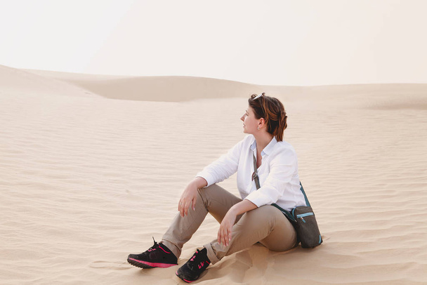Vista lateral de una joven mujer relajada vestida con camisa blanca con jeans beige y zapatillas de deporte mirando hacia otro lado mientras está sentada en la arena en un día soleado.
 - Foto, Imagen