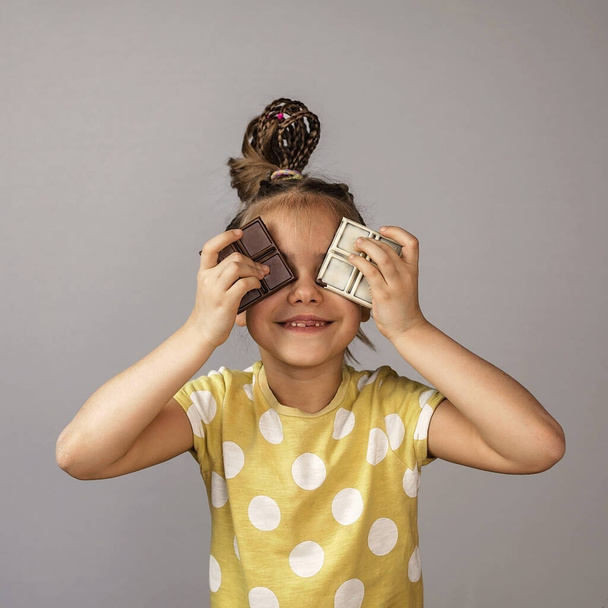 Stijlvol schoolmeisje met zwart-wit chocoladerepen in beide handen voor haar ogen tijdens zoek-en-verstoppertje, gezonde voeding concept, chocolade gezicht, studio schot, wereld chocolade dag - Foto, afbeelding