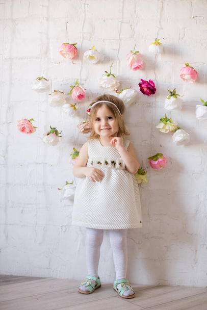 Little cute girl in a beautiful white dress in a spring light studio decorated with flowers - Foto, afbeelding