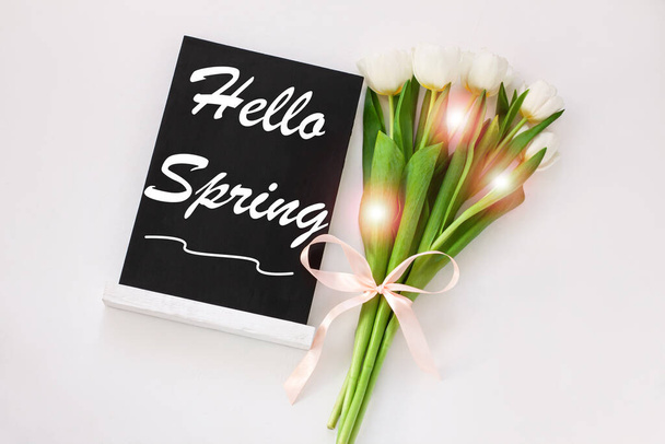 Hello Spring greeting message sign on black chalk board with blooming tulip flowers on white background. Blackboard with text, social media blogs banner. Green leaves, natural floral bloom decoration. - Photo, Image