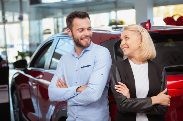 Krásný pár se na sebe vesele usmívá, stojí u svého nového auta v prodejně. Manžel a manželka slaví nákup nového automobilu - Fotografie, Obrázek
